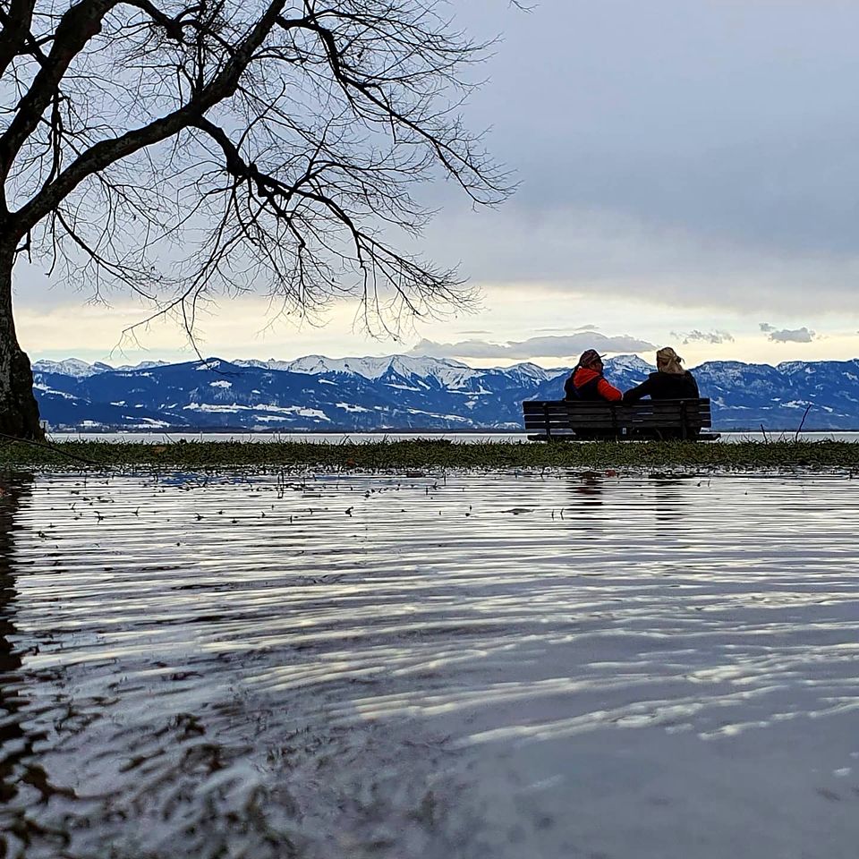 Zwei Spaziergänger auf einer Bank mit Blick auf die verschneiten Alpen